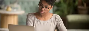 A person wearing glasses sits at a table using a laptop, looking down at the screen while writing on a piece of paper. The setting appears to be a softly lit indoor space with a blurred background, including light-colored furniture and greenery.