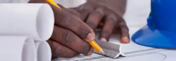 Person writing on a blueprint with pencil beside rolled plans and a blue hard hat