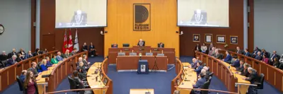A formal meeting is taking place in a large council chamber with Durham Region signage. Attendees are seated in a semi-circle facing the central podium, where a person is speaking. Large screens display the speaker, and flags are visible on the side. The chamber is well-lit with a modern design.