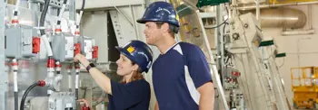 Two workers in safety helmets inspect and operate industrial equipment in a mechanical room