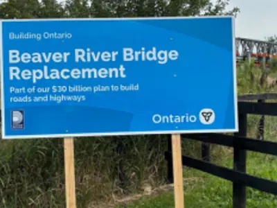 A sign announcing the Beaver River Bridge Replacement, with the temporary Beaver River Bridge in the background.