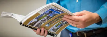 A person in a blue shirt holds and flips through a spiral-bound booklet or catalog featuring colorful photos of buildings or properties. A small round sticker is visible on one page.