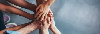 A group of diverse hands placed on top of each other in a gesture of unity against a blurred gray background.