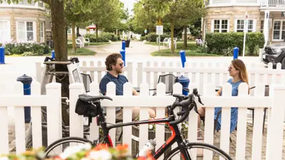 Couple sitting at a bistro table outside a cafe with a bike and flowers in the foreground and a white picket fence.