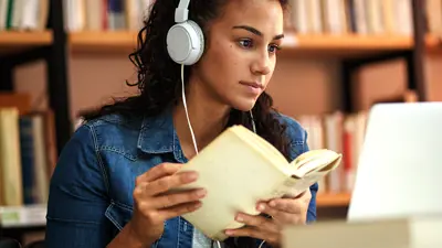 Person wearing over-ear headphones sits at a desk in a library, holding an open book while looking at a laptop. Bookshelves filled with books line the background, and the person’s hands rest on the book as they appear to read and study.
