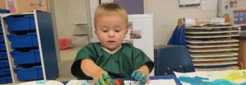 Child in a classroom wearing a green smock finger-paints with bright blue, yellow, and green paint on paper at a table, with art supplies and shelves in the background.